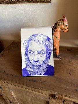 A blue ink portrait drawing of a bearded man displayed on a small easel on a wooden surface, with a decorative wooden horse figurine beside it.