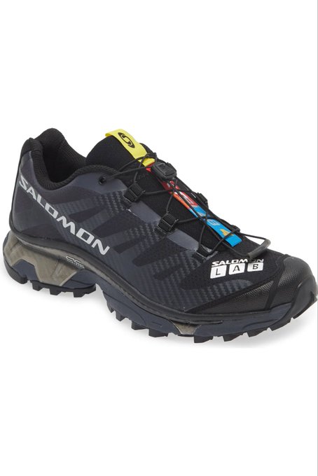 A black Salomon athletic shoe with a textured upper, colorful speckled laces, and a rugged sole is displayed against a white background, with 'SALOMON' branding visible on the side.