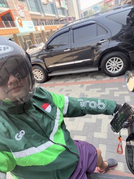 A Grab rider in a green uniform and helmet takes a selfie while checking his phone on a city street, representing the gig economy work referenced in the post about continuing to run Grab today.