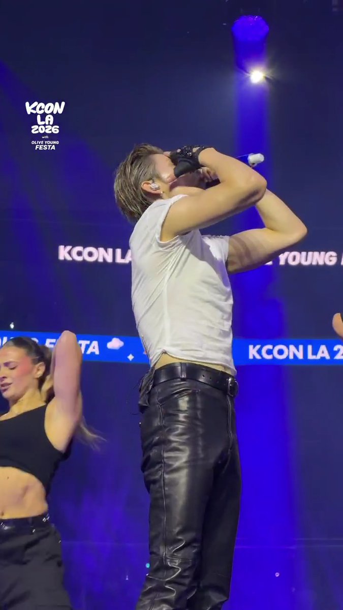 A performer in a white tank top and black leather pants passionately sings into a microphone at the KCON LA 2026 countdown stage, with "KCON LA 2026 with OLIVE YOUNG FESTA" and "KCON LA" visible on the backdrop, capturing Yeonjun of TXT's dynamic Day 2 performance.