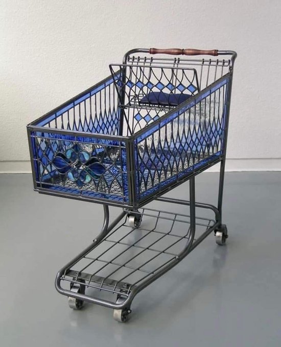A vintage-style metal wire shopping cart features decorative blue stained-glass-like panels with diamond and circular patterns, standing on a gray surface against a white wall.