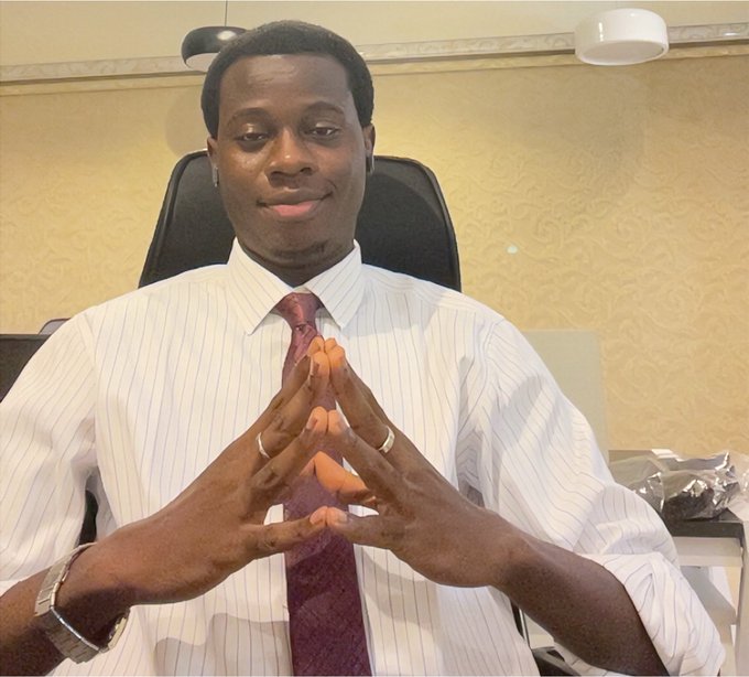 A young man wearing a white striped shirt and a maroon tie sits in a black office chair with his hands pressed together in front of him, wearing rings and a watch.