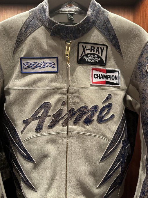 Close-up photograph of a cream-colored leather racing/motorcycle jacket on a hanger, adorned with patches reading 'ZZ-R', 'X-RAY Vanson Performance Products', and 'Champion', with 'Aime' written in stylized dark lettering across the chest.