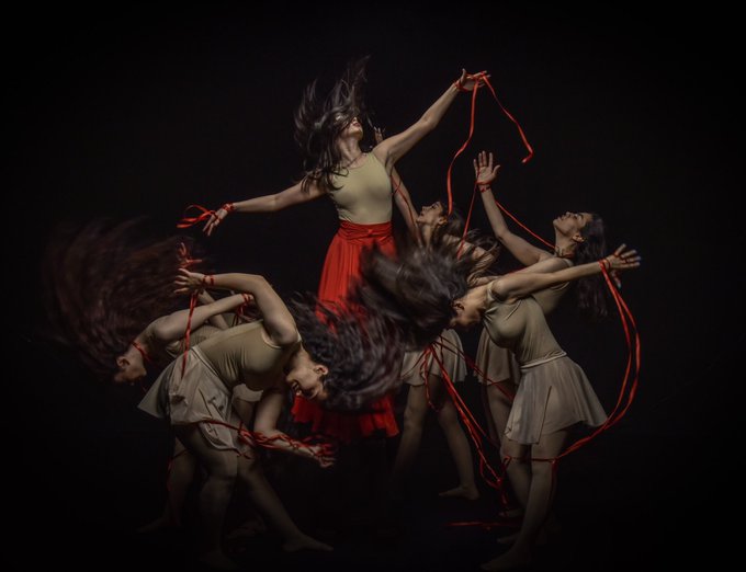 A dynamic dance performance captures multiple dancers in motion, one central figure in a red skirt holding red ribbons while the others swirl around her against a black background.
