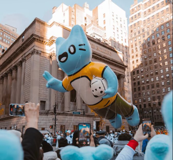 A parade scene featuring a large blue cat balloon in a yellow shirt being paraded down a city street while a crowd takes photos with their phones