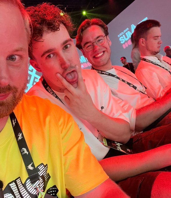 Four men wearing lanyards and casual clothing pose playfully for a selfie at what appears to be the Talent Summit event, with the text "TALENT SUMMIT" visible in red and white on the backdrop. The post humorously suggests their exaggerated, goofy expressions capture the over-the-top excitement of seeing a portfolio reach six figures.