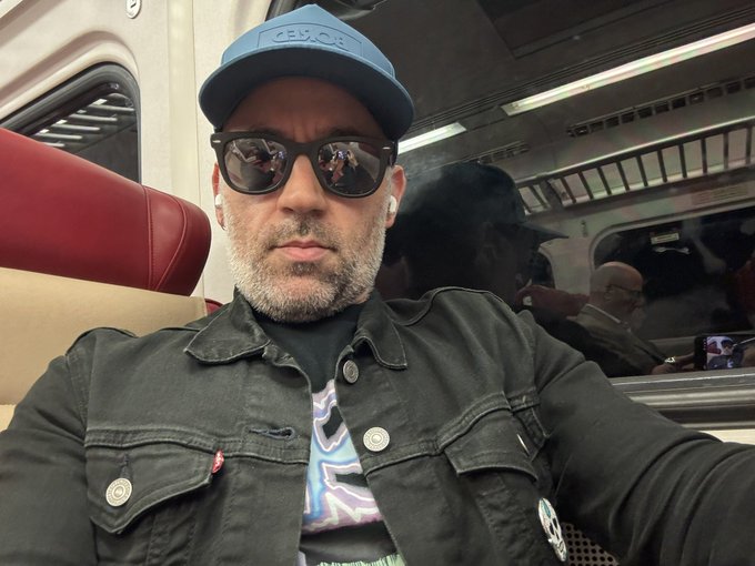 A man with a graying beard wearing sunglasses, earbuds, a blue REV cap, and a dark denim jacket takes a selfie on a train with red seats.