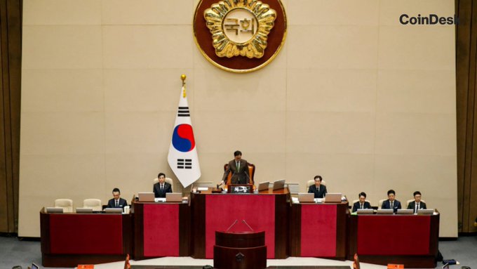 A formal South Korean legislative chamber is shown, featuring the national flag and an ornate emblem above a podium where officials are seated during a session. The image relates to South Korea's upcoming 22% tax on crypto gains above $1,740, set to take effect January 1, 2027. A "CoinDesk" watermark appears in the upper right corner.