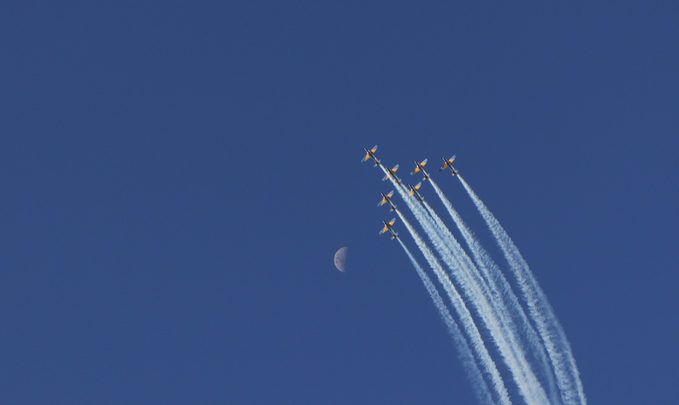 A formation of airplanes flies diagonally across a deep blue sky, leaving long white vapor trails, with a faint crescent moon visible in the background.