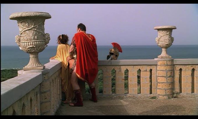 A man in a Roman centurion costume with a red cape stands on a seaside terrace with a woman in yellow robes, looking out over the ocean alongside another figure wearing a plumed helmet. The post's shorting strategy for ETH mirrors the scene's classical "racing toward a cliff edge" motif—boldly betting on a dramatic fall from a precarious elevated position.
