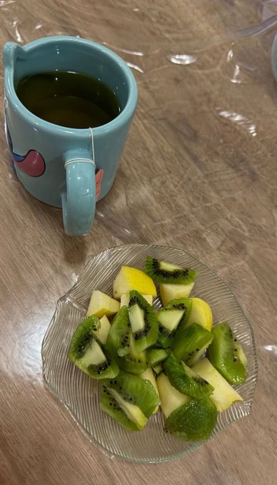 A casual photo of a teal mug containing tea with a tea bag, sitting next to a glass plate filled with sliced kiwi on a wooden table.