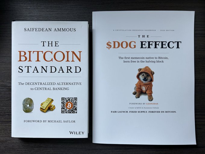 Two books on a dark surface: Saifedean Ammous's 'The Bitcoin Standard' (foreword by Michael Saylor) and 'The $DOG Effect' by Leonidas, a 2026 Cryptolution Research Handbook featuring a Shiba Inu in an orange hoodie.