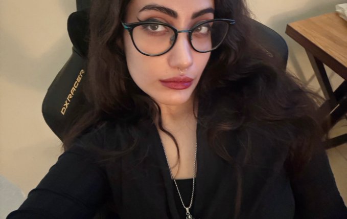 A selfie-style photo of a woman with long dark wavy hair, cat-eye glasses, and a small nose ring, sitting in a DXRacer gaming chair wearing a black top and silver chain necklace.