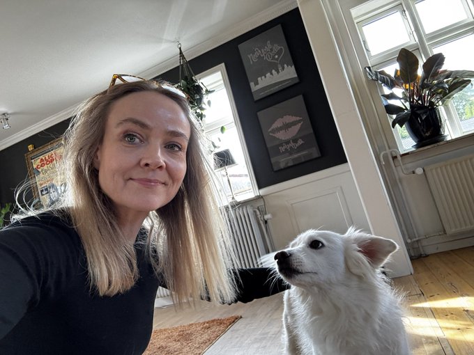 A woman with long blonde hair takes a selfie indoors next to a small white dog, with framed Rolling Stones and Nirvana art prints visible on the dark wall behind them.