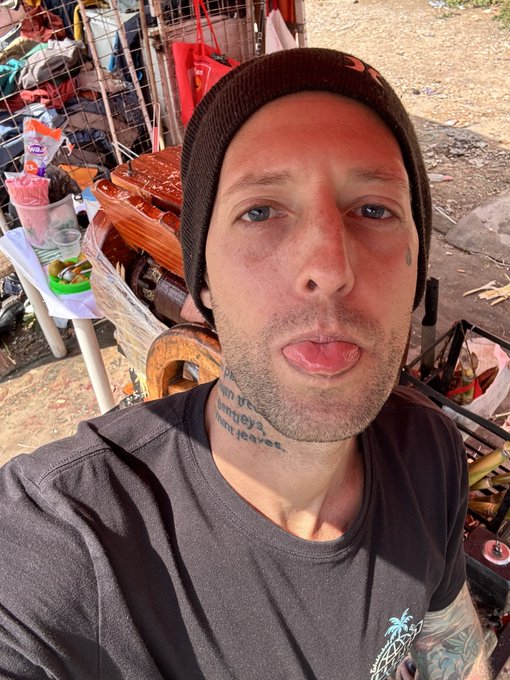 A man in a black beanie sticks out his tongue for a selfie outdoors, surrounded by cluttered storage items. His neck tattoo reads "Monkeys [I] won't leaves" — a playful, carefree vibe matching the cheerful "Good morning" greeting.