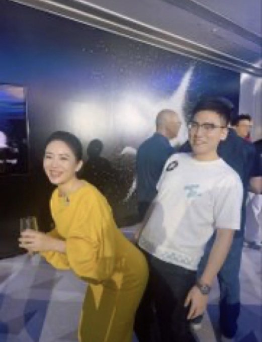 A woman in a yellow dress holding a glass and a man in a white T-shirt with "CZ" printed on it are smiling and posing at a social event, seemingly referencing the playful rivalry mentioned in the post between Star (OKX) and CZ (Binance).