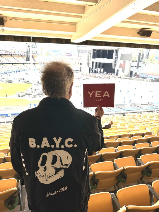 A man viewed from behind wearing a denim jacket with 'B.A.Y.C. Yacht Club' and a skull design, holding up a red sign that reads 'YEA (ㅛㅁ)' while looking toward a stadium field.