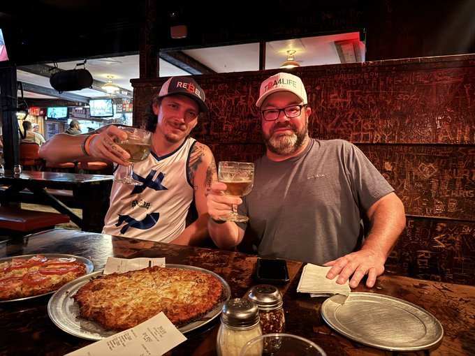 A man and his "Bitcoin Brother" raise their glasses in a toast over a Moose Saloon pizza and beer, celebrating an in-person meetup at the restaurant. Visible text on the wall matches the "Moose Saloon" reference, while printed receipts on the table hint at the National Park trip mentioned in the post.