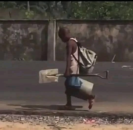 A young boy walks along a dirt road carrying a makeshift toy vehicle constructed from wire, a wooden block, and plastic containers.