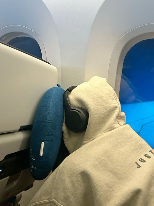A person wearing noise-canceling headphones and a 'JUST' hoodie resting against a blue neck pillow on an airplane seat.