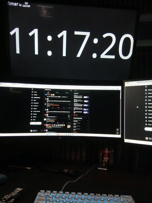 A dimly lit desk setup featuring dual monitors displaying chat applications, a backlit keyboard, and a large timer showing '11:17:20'.