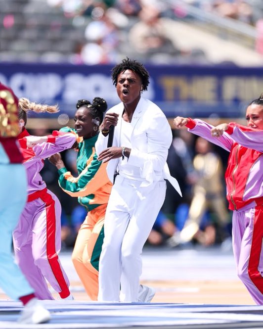 A performer in a white suit singing and dancing energetically with backup dancers in colorful pink, orange, and green tracksuits at what appears to be a stadium event.