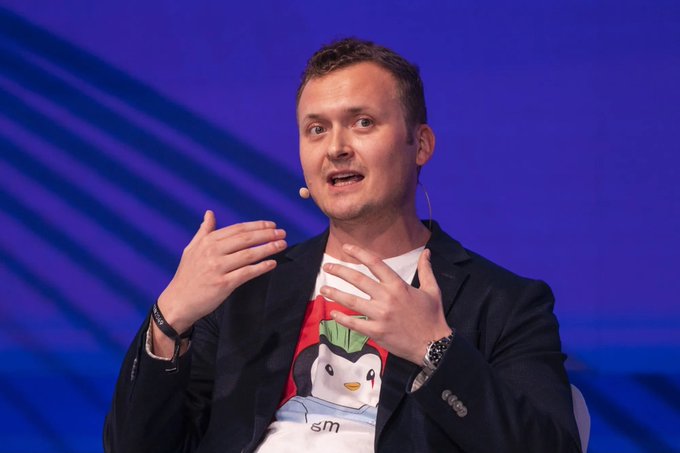 A man with short brown hair speaks on stage against a blue background, wearing a headset microphone, dark blazer, and a graphic t-shirt with partial text reading "sm," while gesturing with both hands raised—appearing to discuss why Bitcoin will never fall below $60K again, as stated in the accompanying post.