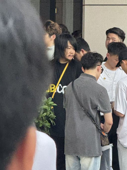 A man with shoulder-length dark hair wearing a black T-shirt with partial white text "BC" visible is standing in a group, seemingly the subject of the casual photo being discussed. Given the context, he appears to be the person being complimented on looking "handsome" with "thick hair" in the meme referenced by the post.