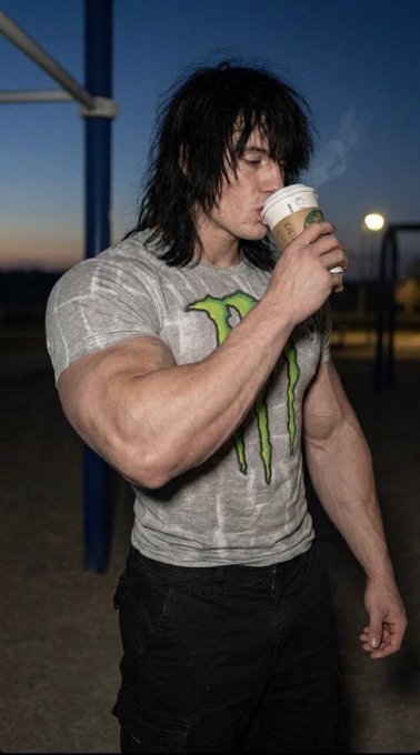 A muscular man with long dark hair drinks from a coffee cup while wearing a gray Monster Energy t-shirt and black jeans at dusk near a basketball court. The image complements the "Drop Wallets" post, suggesting the energy drink brand's sponsorship or promotional content aimed at fitness and active lifestyle enthusiasts.