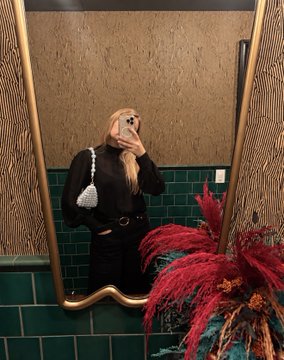 A mirror selfie of a blonde woman holding a phone, wearing a black top and a small white bag, standing in a bathroom with green tile and pink floral decor.