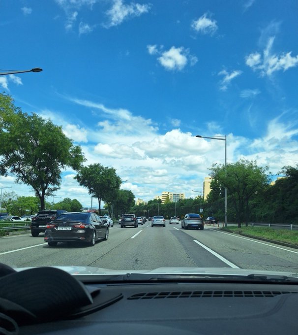 A view from inside a car driving on a multi-lane highway under a bright blue sky with scattered clouds, with a hat visible on the dashboard.