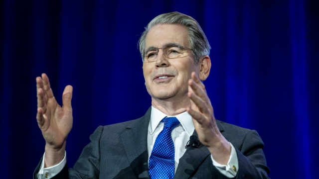 A man in a dark suit with a blue tie and glasses gestures with both hands while speaking against a blue curtain backdrop.