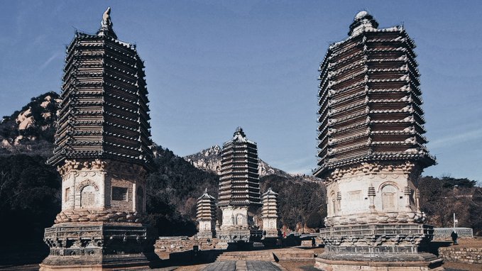 Several tall, ornate stone stupas stand in a courtyard with mountains in the background under a clear blue sky.