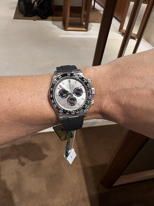 A person's wrist displays a Rolex Daytona Cosmograph watch with a white dial, black ceramic tachymeter bezel, and a dark blue rubber strap, complete with a green price/authenticity tag.