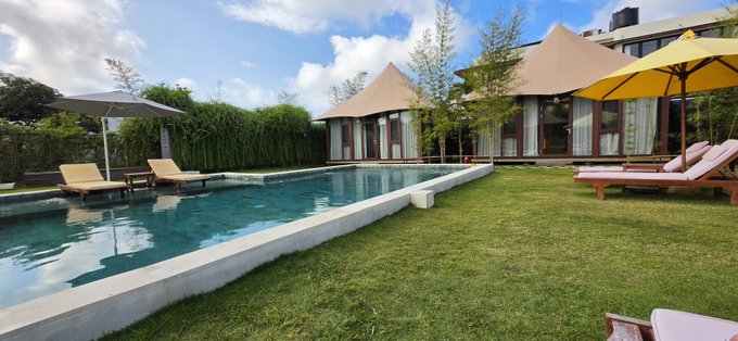 An outdoor resort area featuring a rectangular swimming pool surrounded by a green lawn, lounge chairs with yellow umbrellas, and tent-style cabanas in the background.