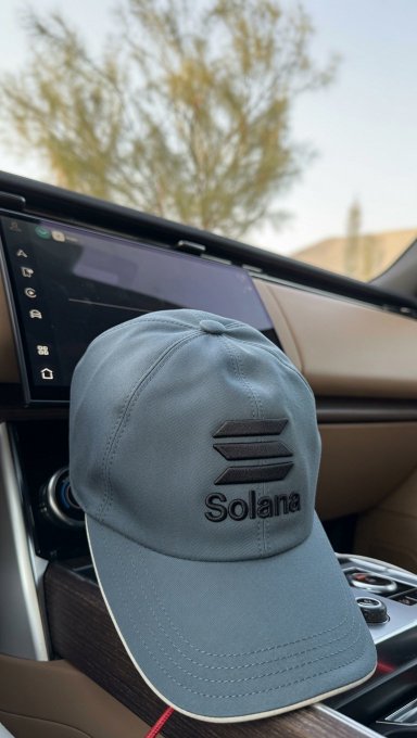 The image features a blue baseball cap with the word "SOLANA" written on it, placed inside of a car's sunroof. The car has a tan interior that matches the color of the hat. The background shows a tree outside the vehicle, and there is also text visible in the image.