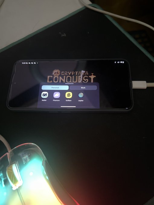 The image shows a cell phone with a gaming app installed, specifically for the game "Conquers". The phone's screen displays the game's interface, which includes various icons related to the game. A mouse can be seen next to the phone on a table or desk, suggesting that it is being used in conjunction with the gaming experience.