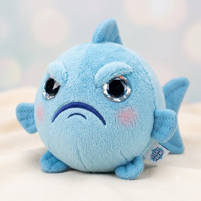 A blue plush toy fish with large crystal/gem eyes, an embroidered frown, pink cheeks, and a 'Fishy Friend' tag, sitting on a light surface.