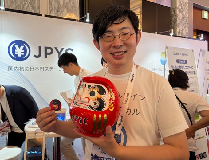 A smiling man wearing glasses holds a red Daruma doll branded with the "JPYC" logo and a small JPYC coin at what appears to be the WebX 2026 conference, where the JPYC booth (Japan's first yen stablecoin) is visible in the background.