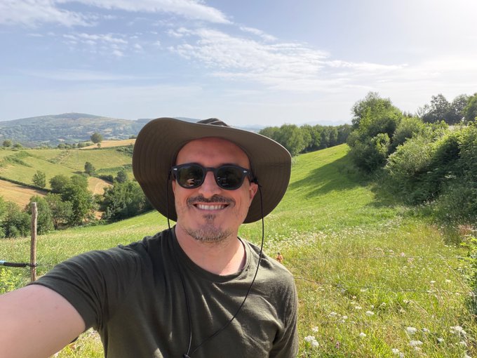 A smiling man wearing a wide-brimmed hat and sunglasses takes a selfie in a lush green meadow with rolling hills in the background.