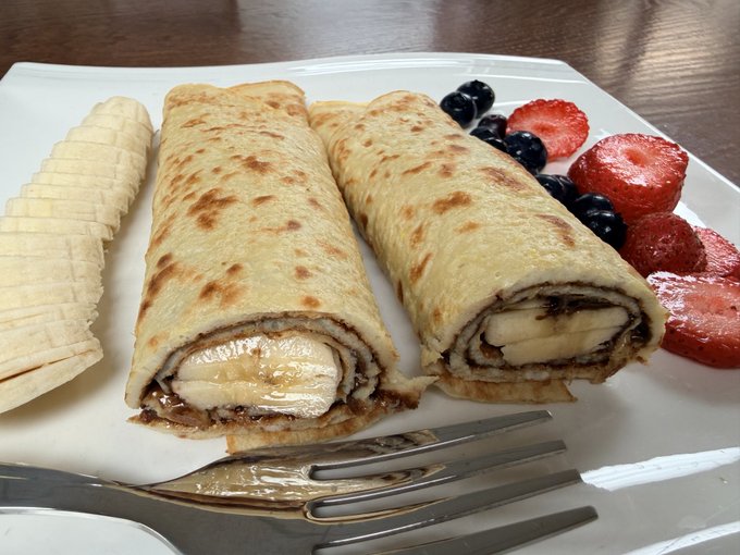 A white plate holds two rolled crepes or pancakes filled with banana and chocolate, served with sliced bananas, blueberries, and strawberries, with a metal fork in the foreground.