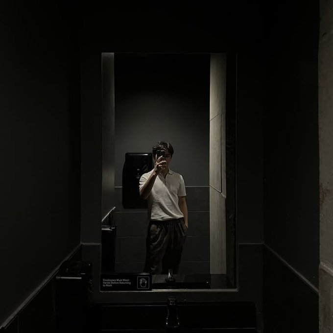 A dark, moody photograph of a person taking a mirror selfie in what appears to be a restroom, with an 'Employee Must Wash Hands Before Returning To Work' sign.