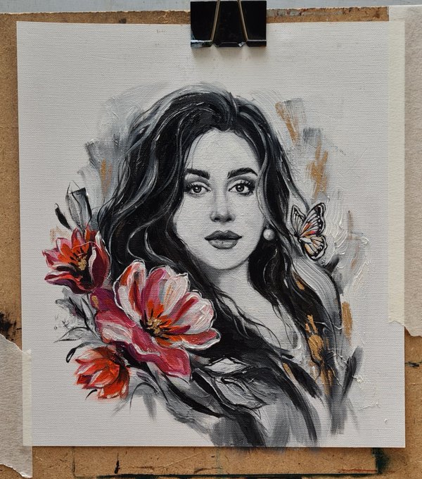 A mixed-media portrait of a woman with long dark hair, rendered in black and white with vibrant red and pink flowers and a small butterfly accent, displayed on a wooden surface held by a black binder clip.