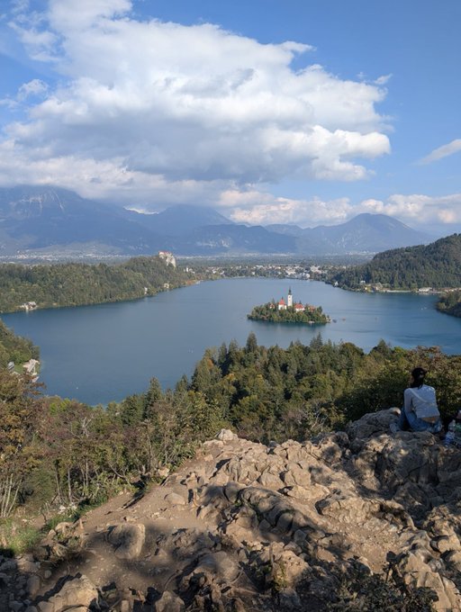 A scenic overlook of a lake with an island containing buildings, surrounded by mountains, with a person sitting on rocky terrain looking out.