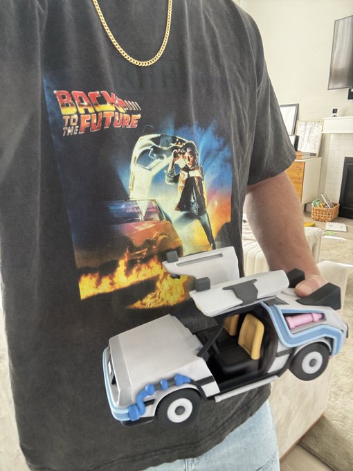 A person wearing a "Back to the Future" KITH t-shirt featuring the iconic DeLorean and Marty McFly holds a custom DeLorean-shaped toy car from the Doodles Toy Factory x DeLorean Labs collaboration, with gull-wing doors open to reveal the interior.