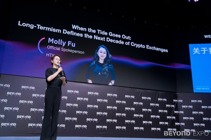 The image features a woman standing on stage at an event, giving a speech or presentation. She appears to be addressing the audience about the crypto industry and its future. The background of the image shows a large screen displaying information related to the crypto world, such as the statement "When the tide goes out, cryptocurrency exchanges go up". This suggests that there might have been a significant event or news related to the crypto market recently.
