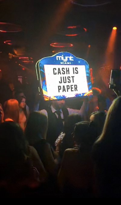 A nightclub scene with someone holding up an illuminated 'mint MIAMI' sign that reads 'CASH IS JUST PAPER' above a crowd under red lighting.