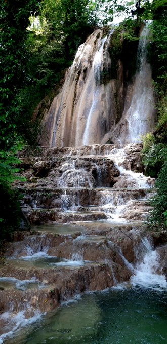 A multi-tiered waterfall cascades down rocky steps surrounded by lush green foliage into a pool below.