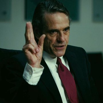 A man in a dark suit and red tie holds up two fingers while speaking.