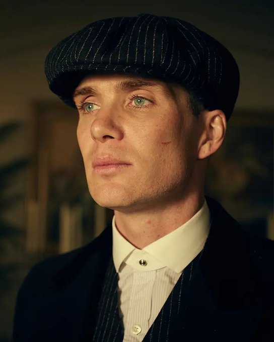 A cinematic close-up of a pale young man with striking blue eyes wearing a pinstripe flat cap and a formal suit with a white shirt, evoking a Peaky Blinders aesthetic.